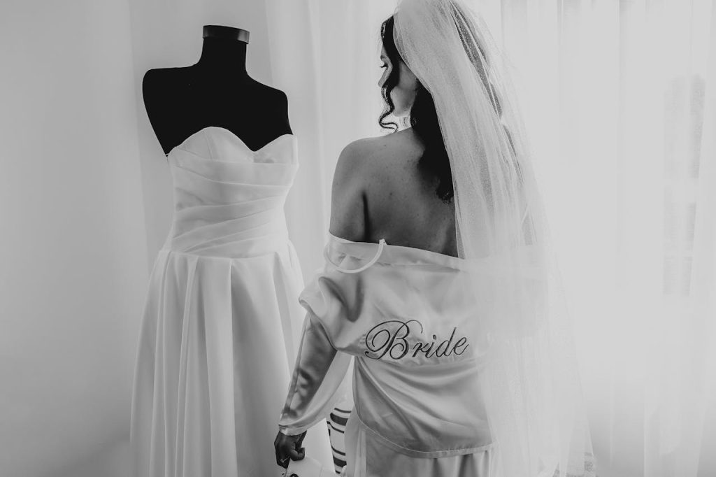 A bride in a robe and veil stands next to her wedding dress, ready for her big day.